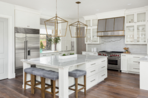 Luxury modern white kitchen featuring marble countertops, brass lantern pendants, gray upholstered bar stools, and stainless steel appliances with hardwood flooring.